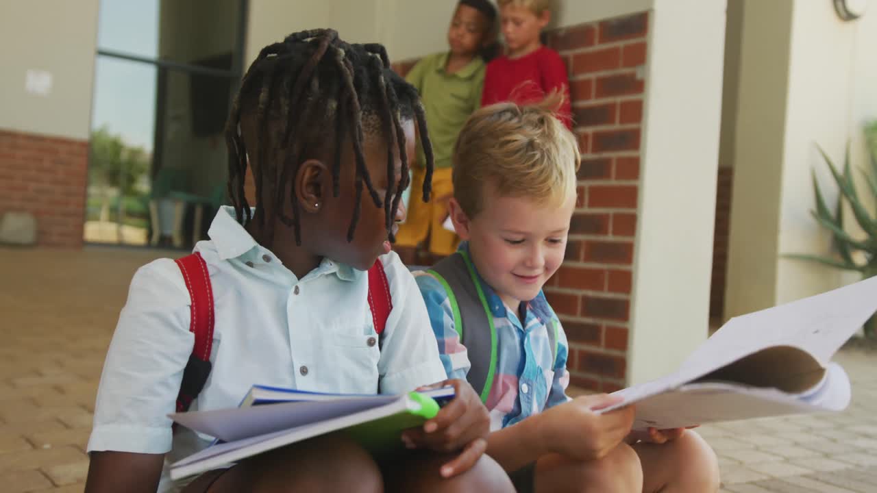 Video of happy diverse boys holding books and talking in front of school