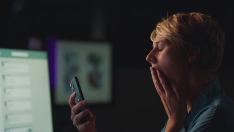 face of tired businesswoman yawning working late in office using mobile phone lit by computer screen