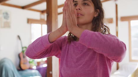 Relaxed-mixed-race-woman-practicing-yoga,-sitting-meditating-in-sunny-cottage-bedroom