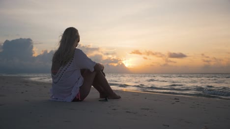 young blonde girl in white shirt sitting alone on a sandy beach watching sunset over the calm ocean, golden hour with blue cloudy sky