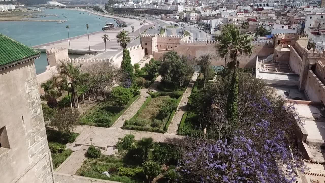 Smuk droneoptagelse med udsigt over de andalusiske haver inden for den historiske Kasbah af Udayas. Viser de befæstede mure, grøntflisede tårne, blomstrende jacarandatræer og flodbaggrund