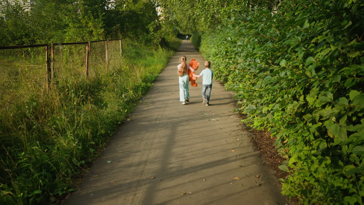 Rear view of cheerful children walking side by side on quiet path with girl holding teddy bear while boy walks beside her. Path is bordered by lush greenery and metal fence under warm sunlight