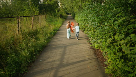 rear view of cheerful children walking side by side on quiet path with girl holding teddy bear while boy walks beside her. path is bordered by lush greenery and metal fence under warm sunlight
