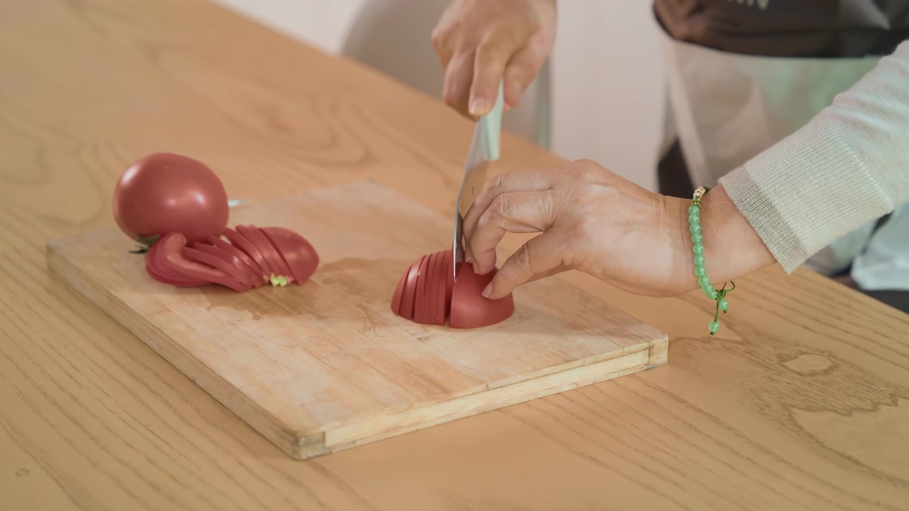 Elderly woman hand preparing healthy meal with fresh vegetables, grandmother wearing an apron chopping slicing tomatoes on wooden cutting board in modern home kitchen, food recipe, meal preparation