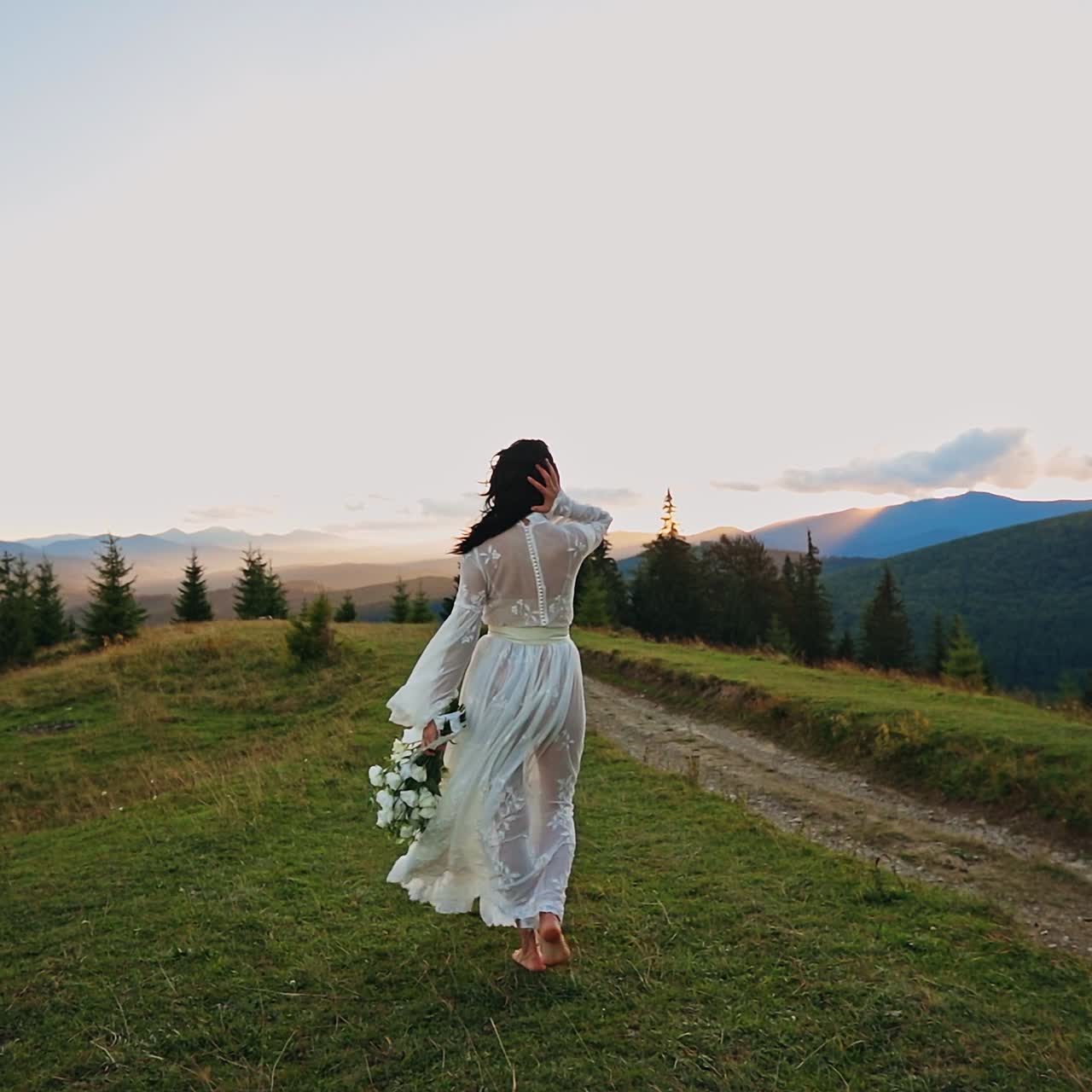 Barefoot lady in transparent white dress walks in the hills. Long-haired woman holding white flowers in her hand. Mountainous scenery with fir-trees at backdrop