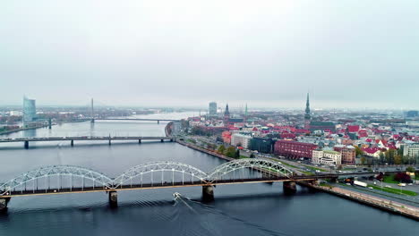 Toma-Aérea-De-Drones-Volando-Sobre-Los-Puentes-Sobre-El-Río-Daugava-Con-El-Casco-Antiguo-De-Riga-En-El-Fondo-En-Un-Día-Nublado-En-Riga,-Letonia