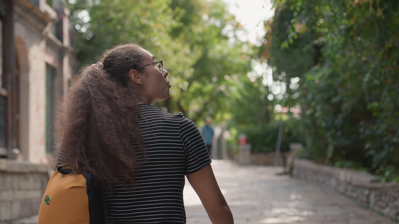 Joven mujer negra caminando por un callejón adoquinado con una mochila amarilla, mirando hacia la copa de los árboles. Postura relajada y expresión curiosa, con vestido de rayas y cola de caballo, mientras la luz del sol de la tarde se filtra.