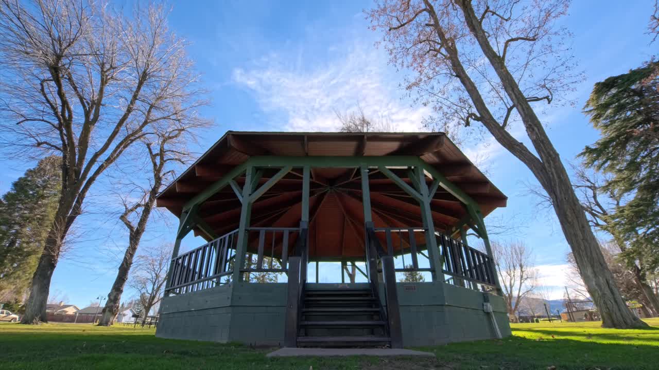 En statisk timelapse, der viser en musikpavillon i en fredfyldt, offentlig park, mens skyerne langsomt driver hen over den blå himmel. Græs og træer indrammer pavillonen og skaber en rolig og reflekterende udendørsscene.