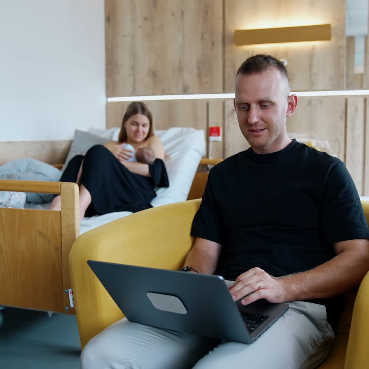 Busy Caucasian man sits in armchair working on laptop. Woman in bed at backdrop breastfeeding her baby