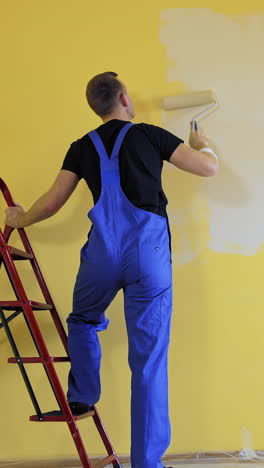 craftsman painting walls near the building ladder. professional painter in blue overalls doing a renovation in the room with a paint roller. rear view. vertical video