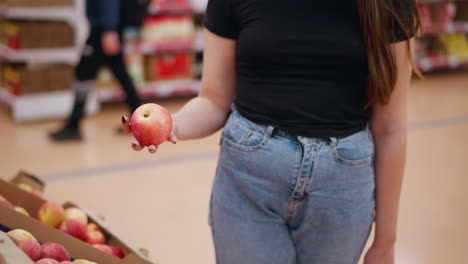 female shopper in grocery store holding shopping bag with left hand, picks red apple from display, observing it, someone passing by in blurred background in vibrant store setting