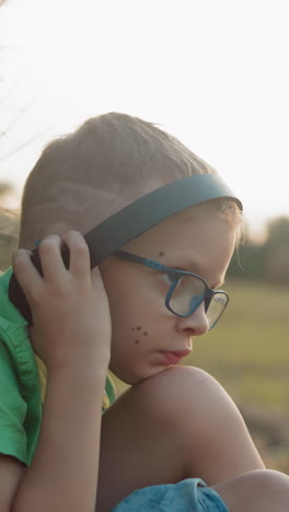 vista lateral de un niño pequeño sentado, ajustando sus auriculares, descansando contra una bala de heno con manchas en su mejilla en un entorno rural sereno