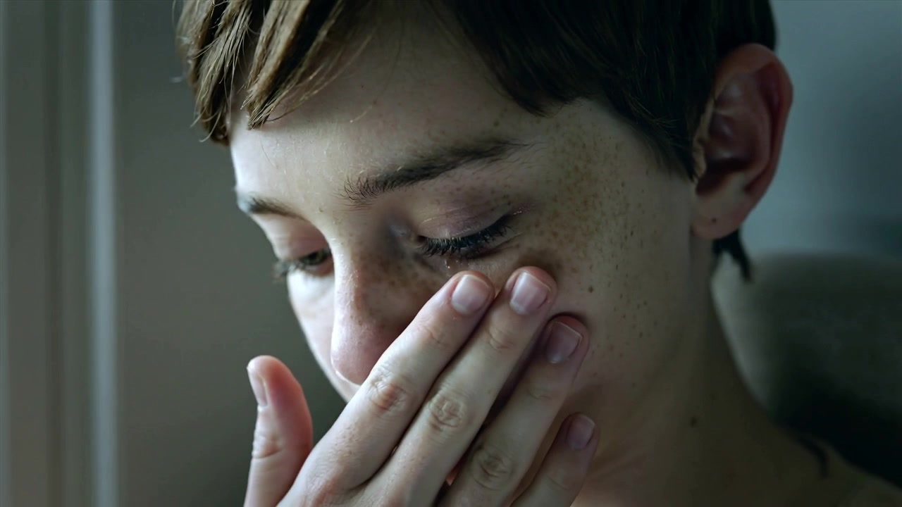 Tear forming, teen lowering head, wiping tear tracks beside home window, ringed hand touching cheek