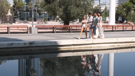 diverse friends talking while walking on footpath by reflecting pool in city during sunny day