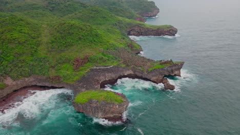 Aerial-footage-of-seascape-with-view-of-big-wave-hits-coastline-of-an-island-that-overgrown-with-dense-forest