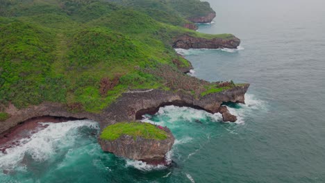 aerial footage of seascape with view of big wave hits coastline of an island that overgrown with dense forest