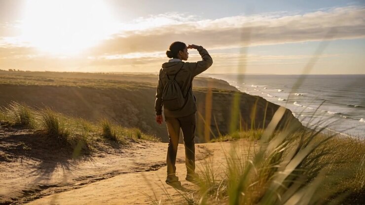 Walking hooded hiker on coastal trail, shielding eyes from low sun and scanning coast with backpack