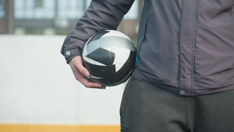 primer plano de la mano sosteniendo una pelota de fútbol en blanco y negro con enfoque en la mano del jugador, mostrando ropa atlética y parte del cuerpo, fondo urbano con edificios y deportes al aire libre