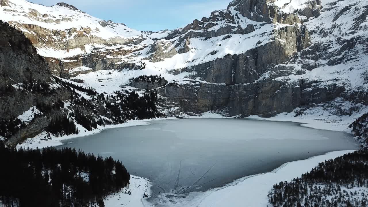 Drone captures a cinematic aerial view of the frozen Oeschinensee, surrounded by snow-covered mountains and forests