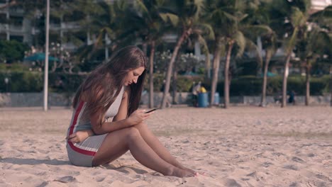 mujer joven descalza en textos grises en el teléfono sentada en la arena