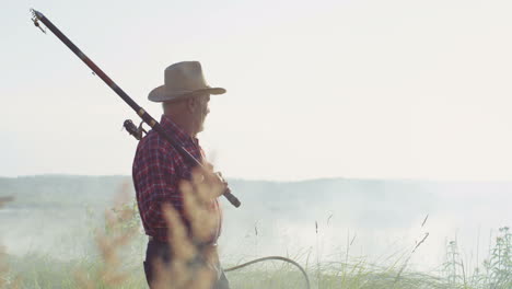 Senior-fisherman-in-a-hat-walking-on-the-lake-shore-with-a-rod-and-equipment-for-fishing-on-a-cloudy-morning