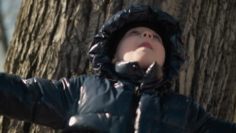 a young boy stands with his back against a large tree trunk, looking upward with his arms fully outstretched, as if embracing the world, he is dressed in a shiny black jacket, jeans, and boots