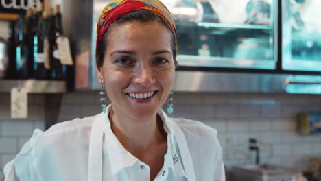 Mujer-Detrás-Del-Mostrador-En-Una-Tienda-De-Delicatessen-Sonriendo-A-La-Cámara