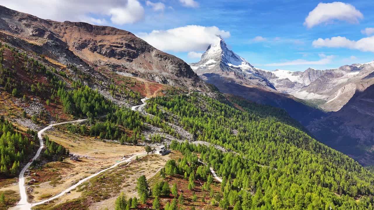 Slow aerial push in toward Switzerland’s iconic Matterhorn peak above Zermatt, highlighting green pine forests, rocky alpine slopes, and serpentine mountain trails stretching toward the snowy summit