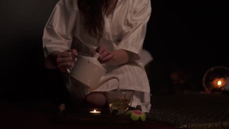 beautiful brunette in bathrobe taking teapot and pouring tea in cup drinking herbal tea at the spa in slow motion. thai tea