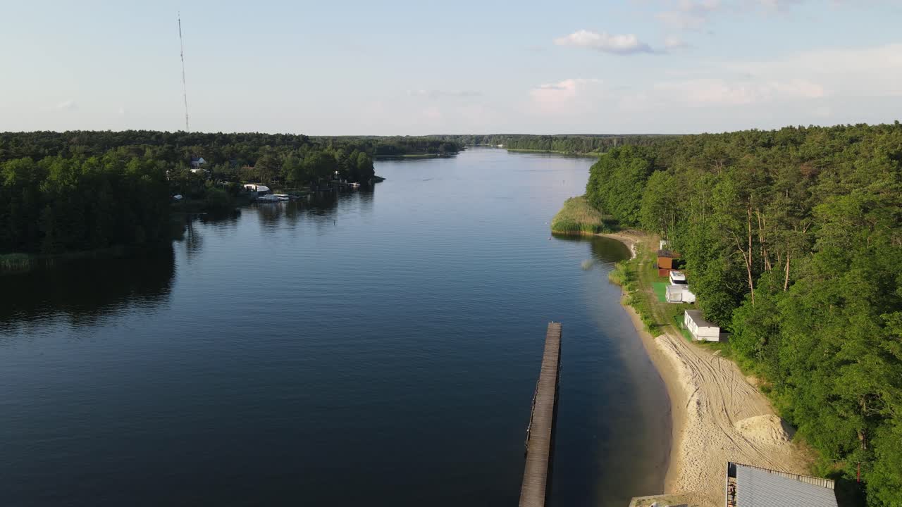 Aerial drone shot descending over a calm forest lake, revealing wooden piers, sandy beach, moored boats and dense green trees under a clear blue sky.