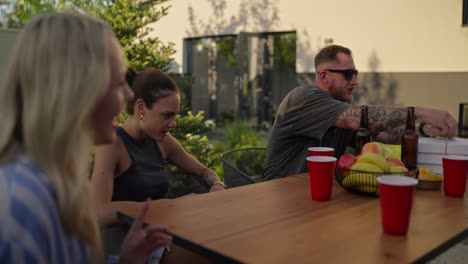 confident guy in sunglasses and a gray t-shirt opens boxes of pizza during a shared lunch at a table in the courtyard of a country house