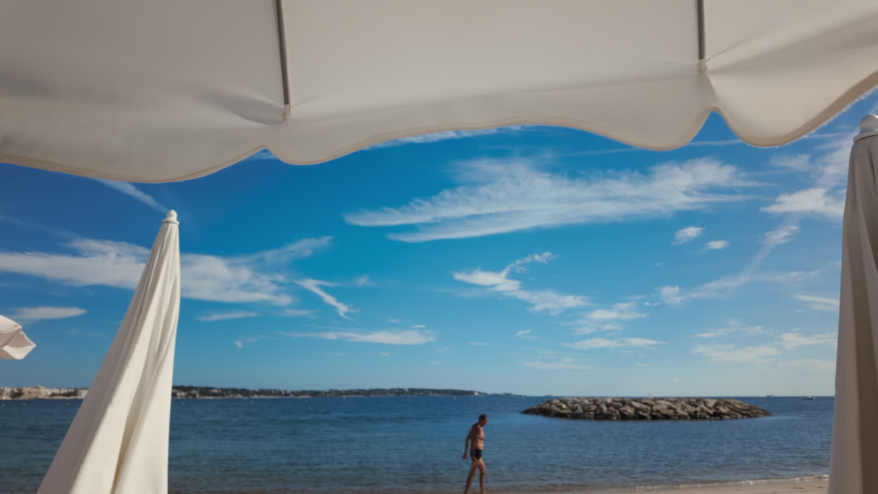 Wide shot of a man walking along the beach under large white umbrellas with a clear blue sky in the background