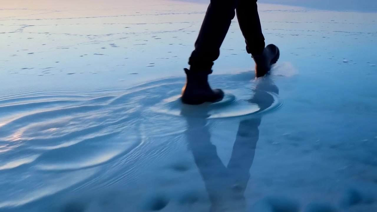 Close-up of lower legs wearing dark waterproof boots stepping through shallow wet sand at low tide, front foot pressing into glossy surface while rear foot lifts, leaving ripples, reflections and
