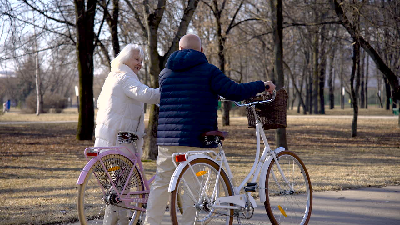 Sett bakfra av et eldre par som holder sykler mens de går og snakker i parken på en vinterdag