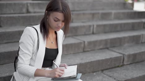 Mujer-De-Negocios-Sentada-En-Los-Escalones-De-La-Ciudad-Escribiendo-Notas-En-Un-Espacio-De-Copia-Láctea