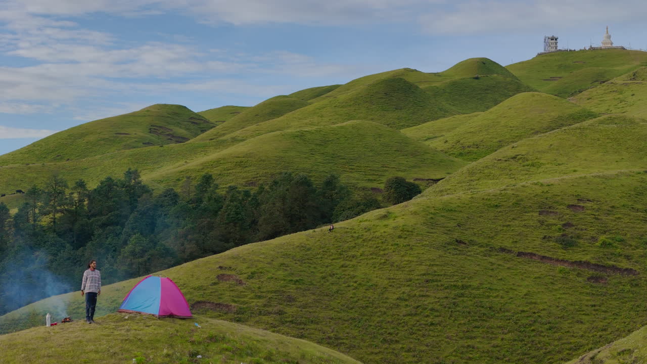 트레커는 티베트 국경 근처에 있는 네팔 도라카의 잔잔한 푸른 언덕에서 캠핑하는 동안 천막에서 나와 화염에 앉아 자연에서 평화롭고 성공적인 솔로 여행을 즐기고 있습니다.