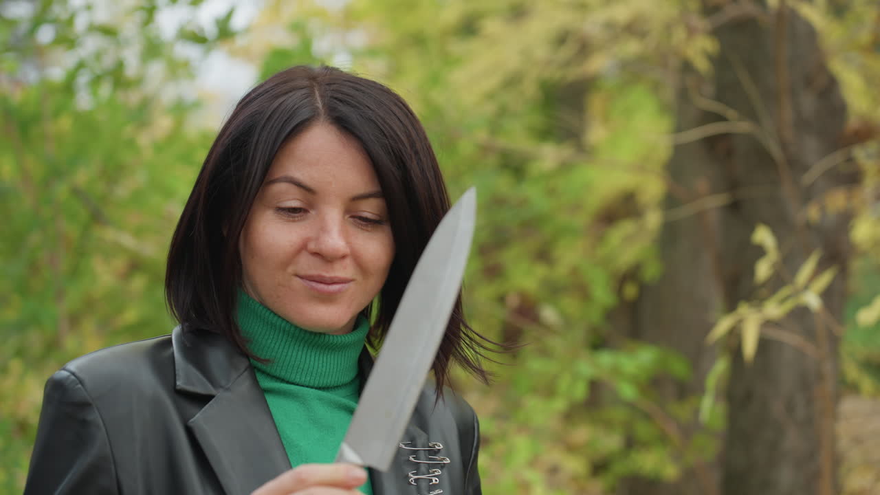 Mulher na floresta segurando uma faca grande, jaqueta de couro e gola rolê verde visíveis, inspecionando a lâmina perto de uma chaleira de acampamento, folhagem de outono e troncos musgosos, olhar confiante e expressão focada, cinematográfico