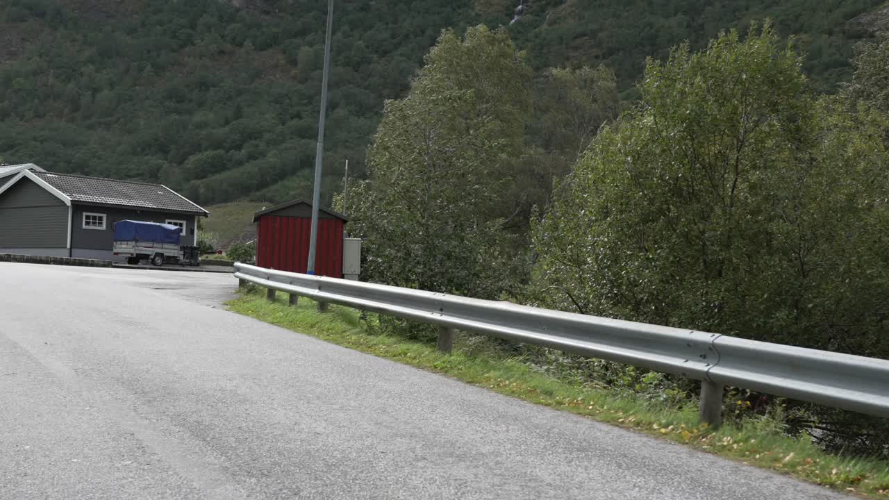 Grey house with white trim and trailer beside forested hillside in Flåm, Norway—quiet residential setup with elevated platform and natural backdrop, shot from a car