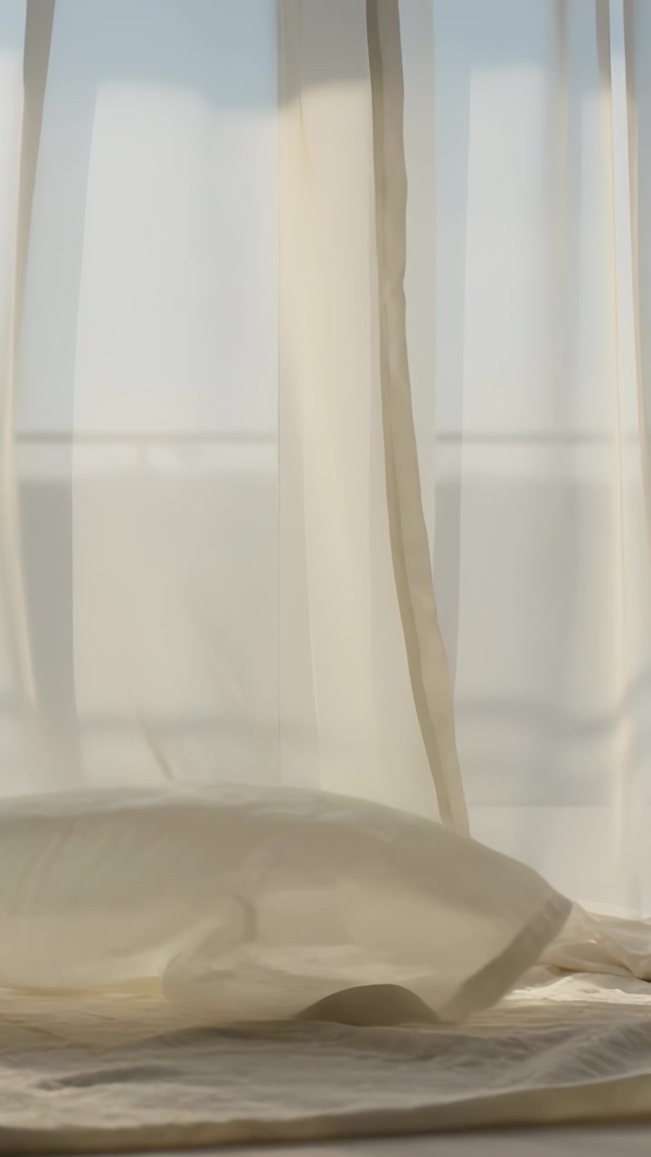 Vertical video: Breeze lifting then settling thin drape and pillow at window sun casting pane shade