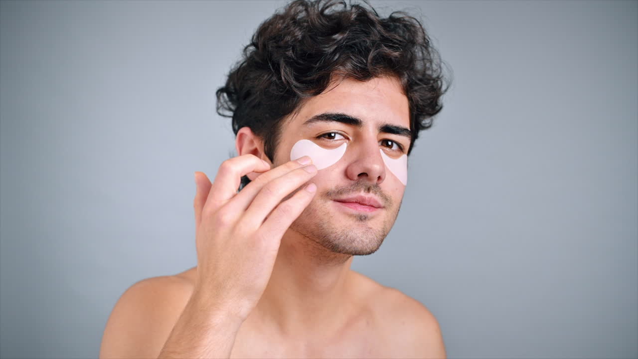 A young caucasian man with stubble beard placing patches under his eyes, looking into the camera