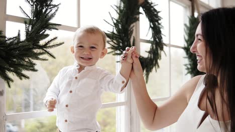 Una-Joven-Madre-Sonriente-Vestida-De-Blanco-Sostiene-La-Mano-De-Su-Hijo-Mientras-él-Camina-Por-El-Alféizar-De-La-Ventana-Decorado-Con-Navidad