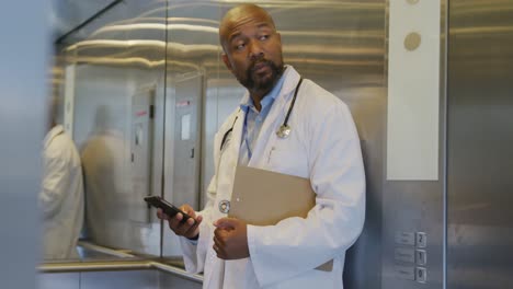 African-american-male-doctor-leaving-elevator-and-using-smartphone-at-hospital