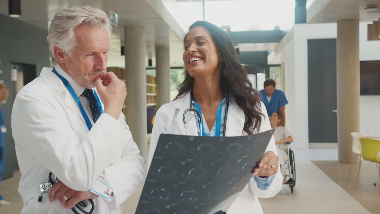 Doctor Wearing White Coat Discussing Patient Scan With Female Colleague In Busy Hospital