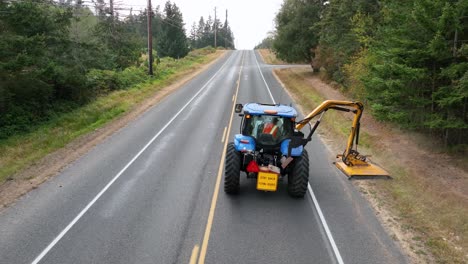 toma aérea apretada siguiendo a un tractor usando una segadora de mayales para nivelar las malezas cubiertas de maleza al lado de la carretera rural