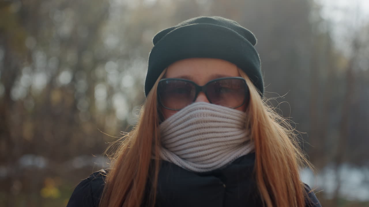 Valkoihoinen nainen puistossa, kasvot peitettynä. Punaiset hiukset pilkistävät neulepipon alta, huivi peittää kasvojen alaosaa. Aurinkolasit peittävät silmät. Yllään musta pipo, reppu ja toppatakki, nainen kävelee metsäpolkua pitkin ja kääntyy.