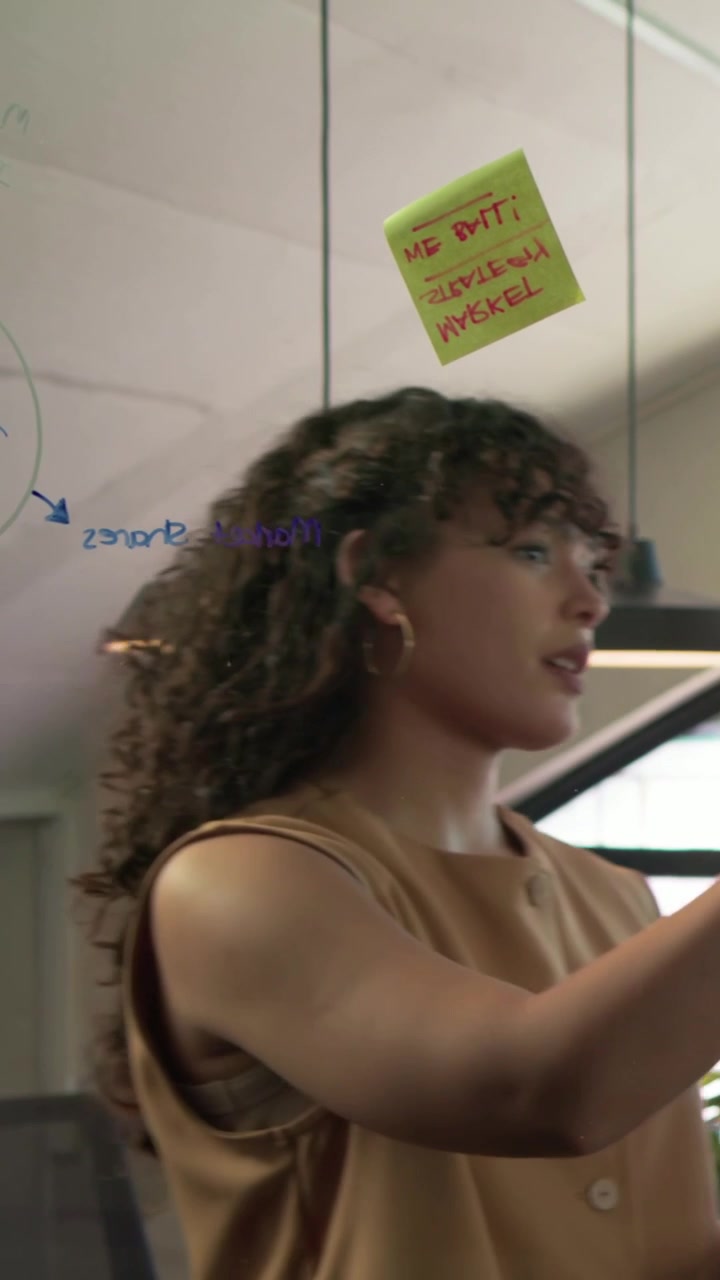 Vertical video: Receiving green notes woman in beige top placing, writing on glass wall in meeting