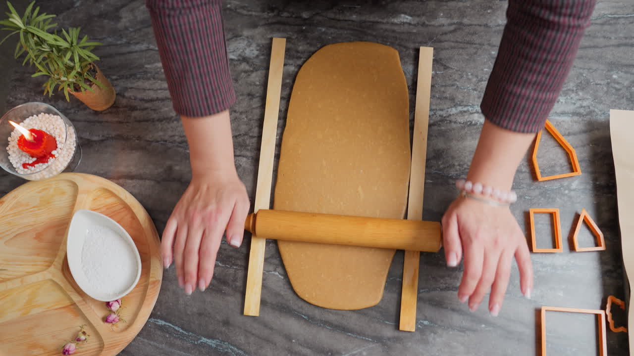 vista aérea de una mujer rodando masa con un rodillo de madera en la encimera, manos presionando firmemente, pulsera de cuentas rosas en la muñeca, rodeada de herramientas de horneado, cortadores de galletas, bandeja de madera y harina