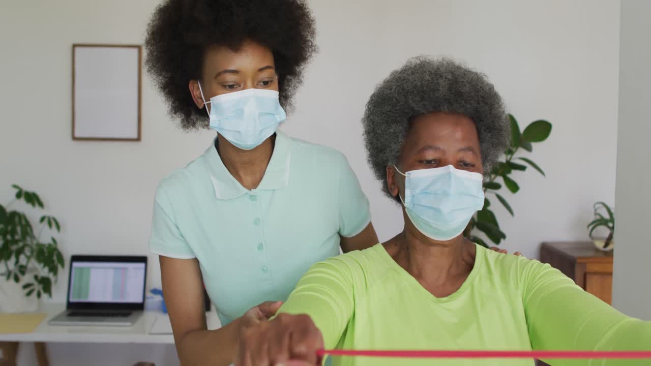 African american female physiotherapist wearing face mask helping senior female patient exercise