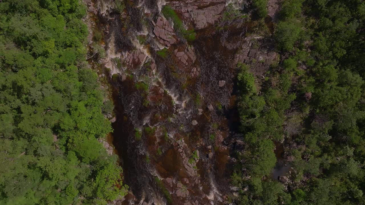 Dark water flowing through rocks and lush vegetation at Cachoeira dos Saltos waterfall in Chapada dos Veadeiros National Park, Goiás, Brazil, on a sunny day, top down drone shot