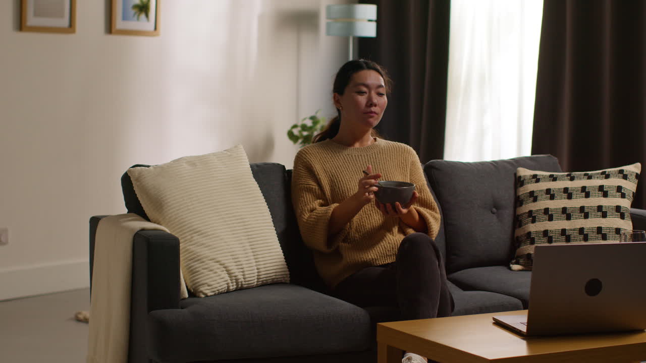 mujer en casa sentada en el sofá comiendo una comida saludable vegetariana o vegana de ensalada tabbouleh con un vaso de vino viendo el programa en la computadora portátil 1